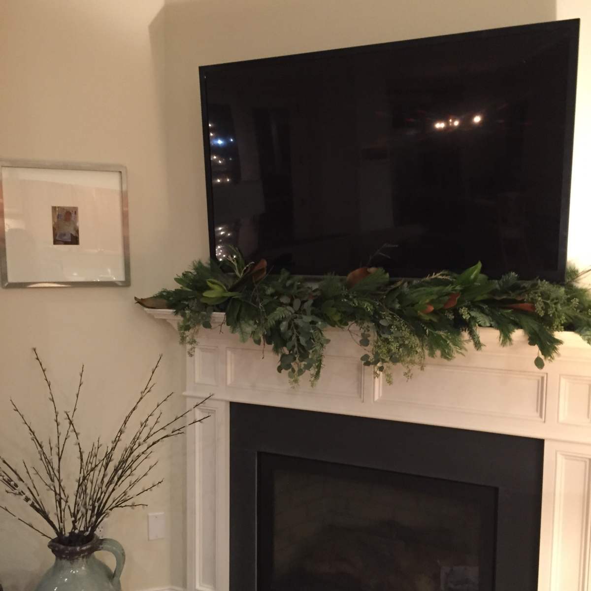 A flat-screen television is mounted above a mantel adorned with greenery, above a black fireplace with a simple vase and branches nearby.