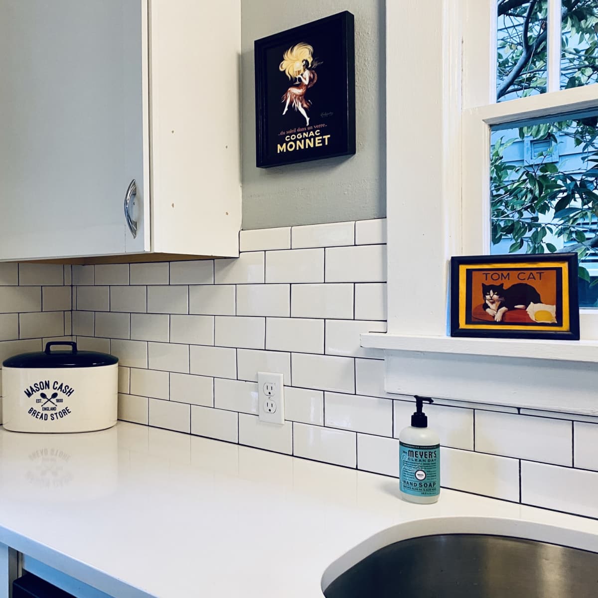A kitchen corner with white tiled walls, a countertop, various framed art pieces, a black and white canister, and a hand soap dispenser.