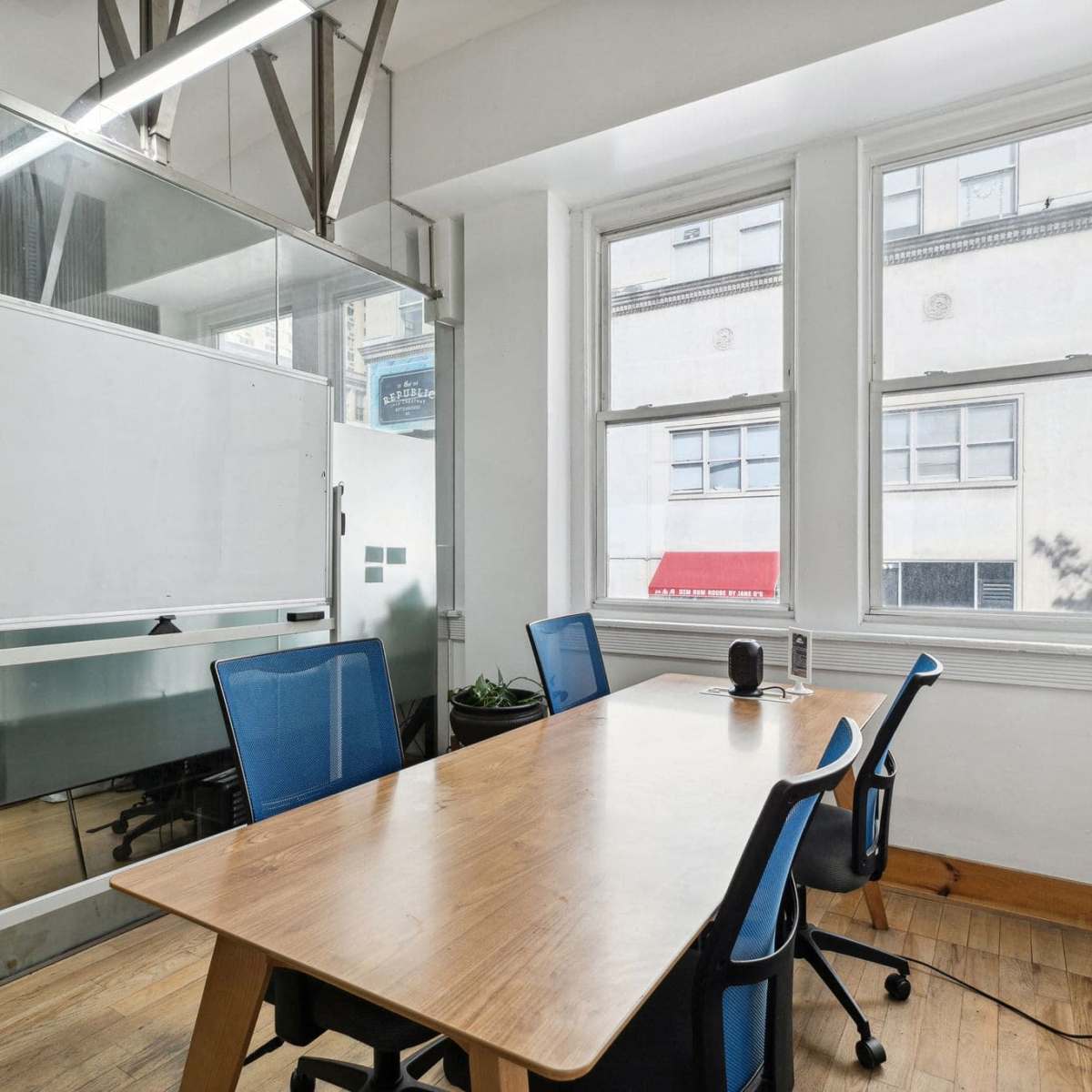 A small conference room features a wooden table surrounded by four blue chairs, with a whiteboard and large windows providing natural light.