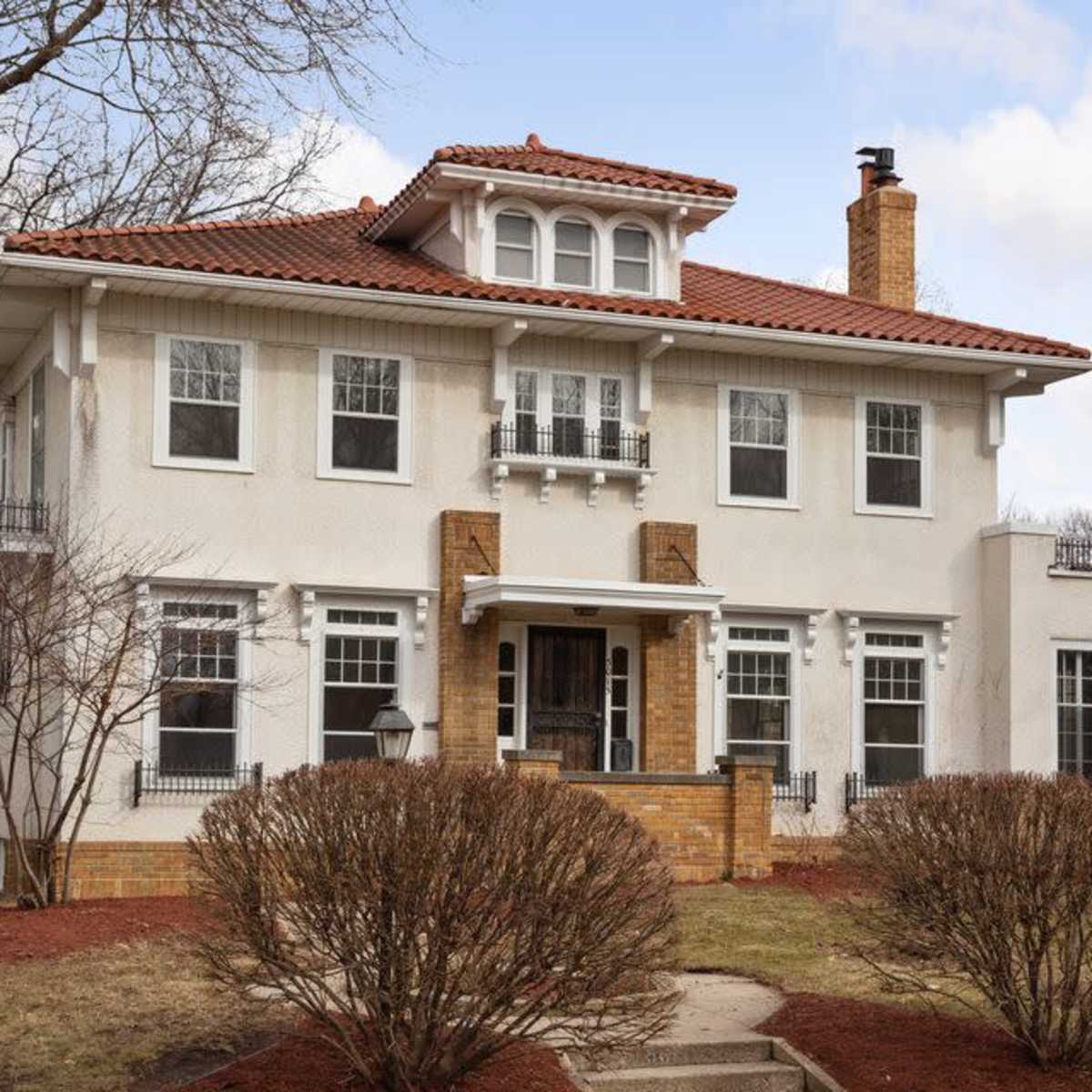 A two-story, cream-colored house with a red tile roof and a brick chimney, surrounded by manicured shrubs and a clear sky.