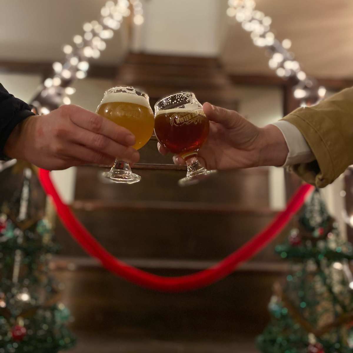 Two hands clink glasses of beer on a staircase adorned with twinkling lights and Christmas decorations.
