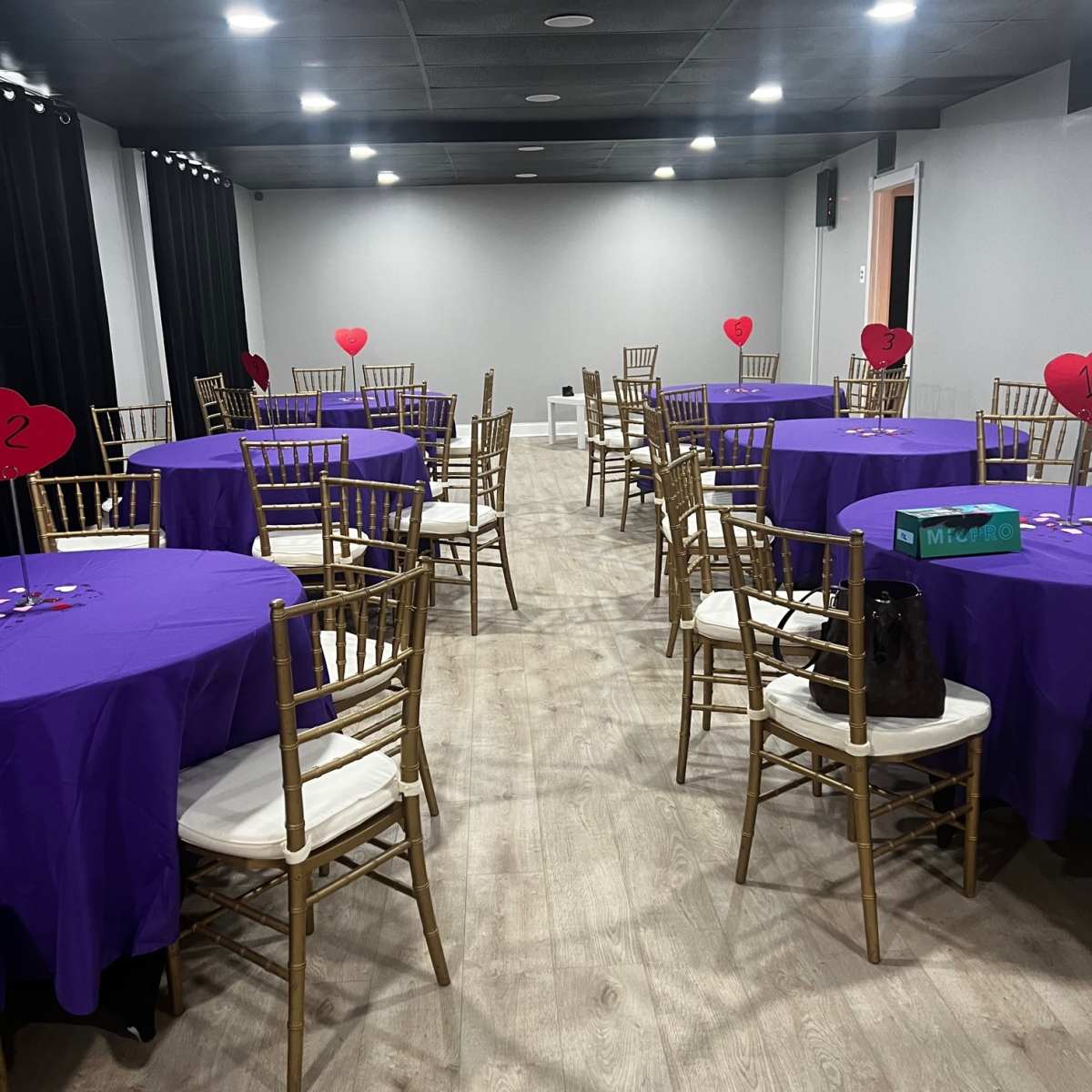 A banquet hall set up with several round tables covered in purple cloths, each surrounded by gold chairs and decorated with heart-shaped centerpieces.