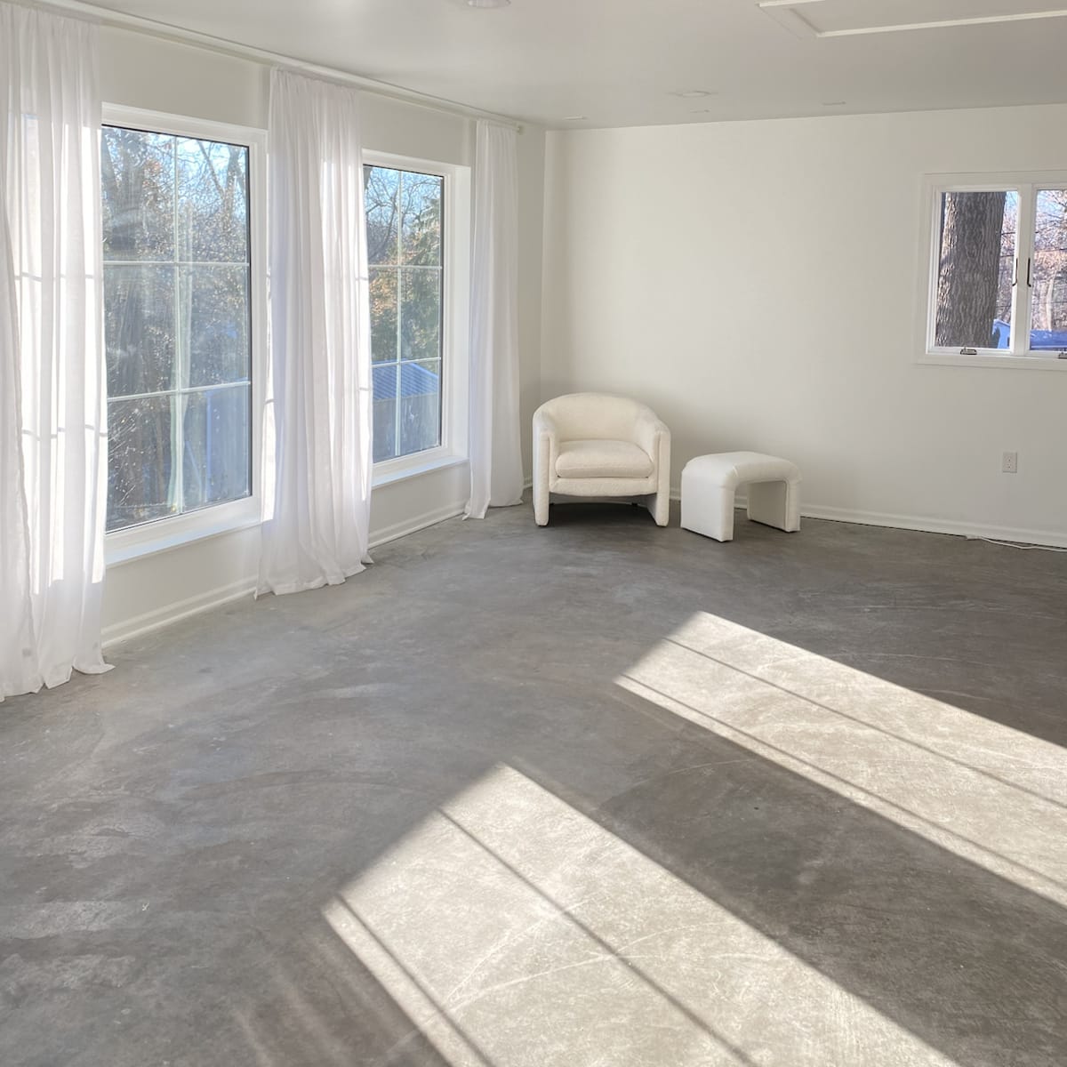 The room features large windows with sheer curtains, a white armchair, and a small stool on a bare concrete floor.