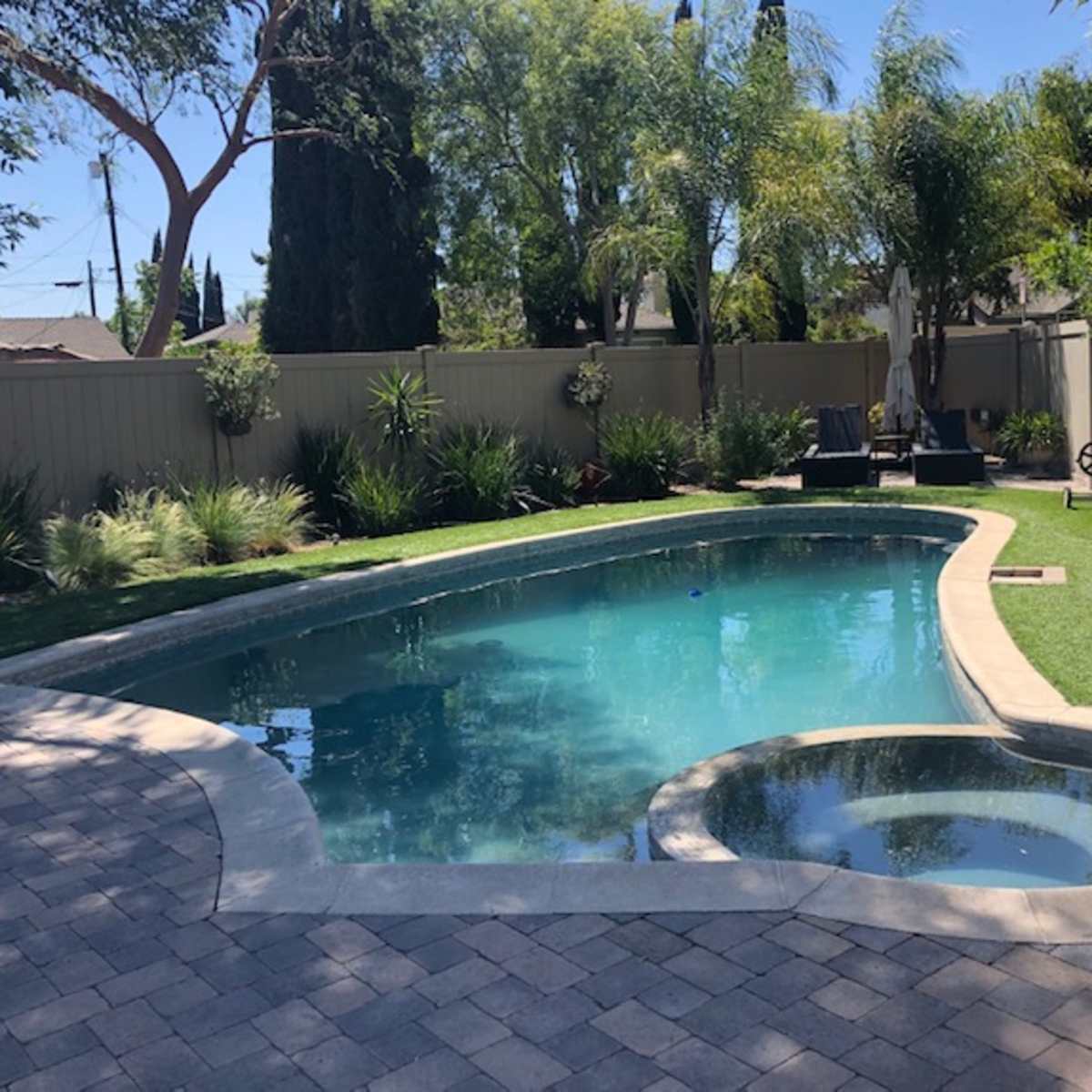 A landscaped backyard features a clear swimming pool with a spa, surrounded by greenery and paved stone.
