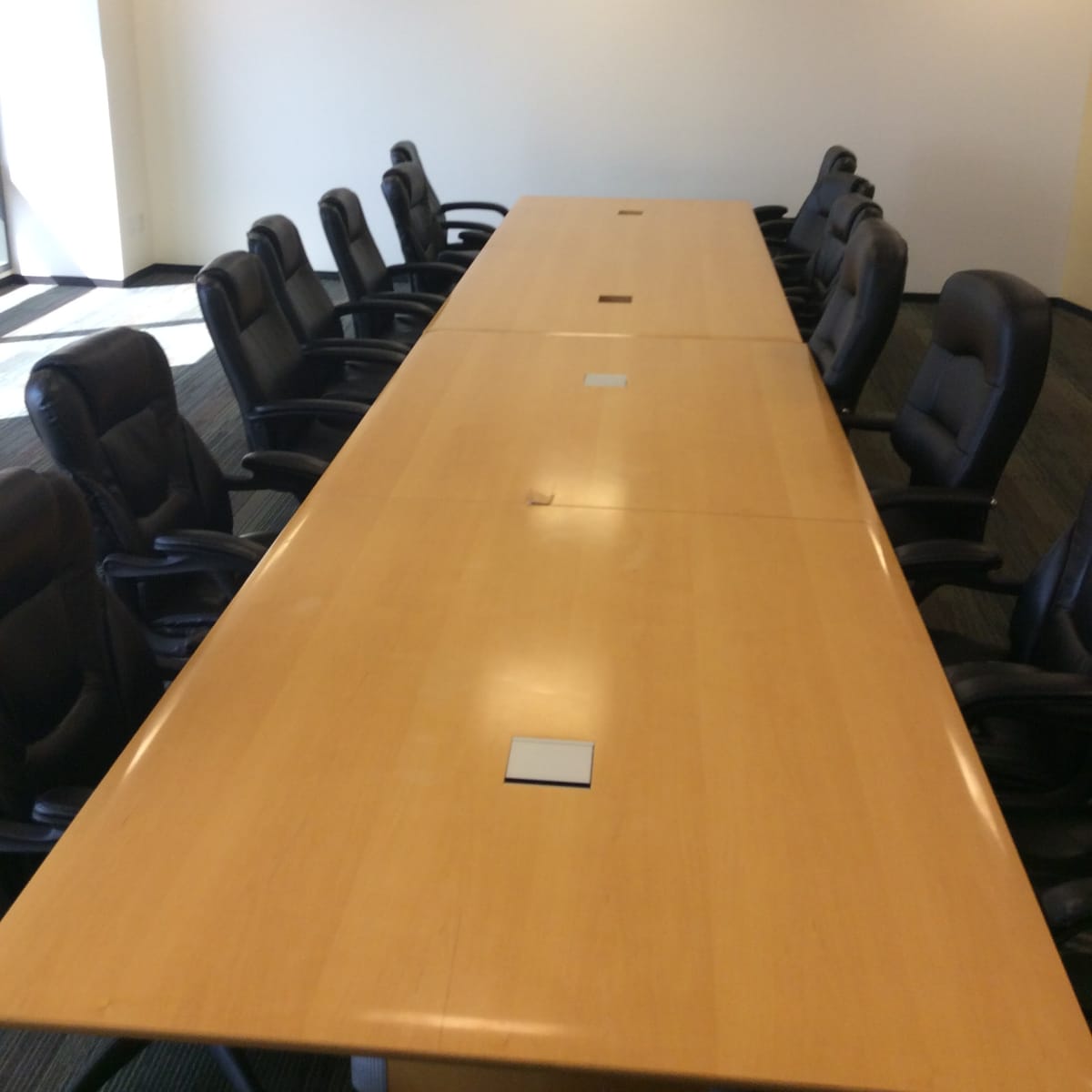 A long wooden conference table with black leather chairs is set up in a brightly lit meeting room.