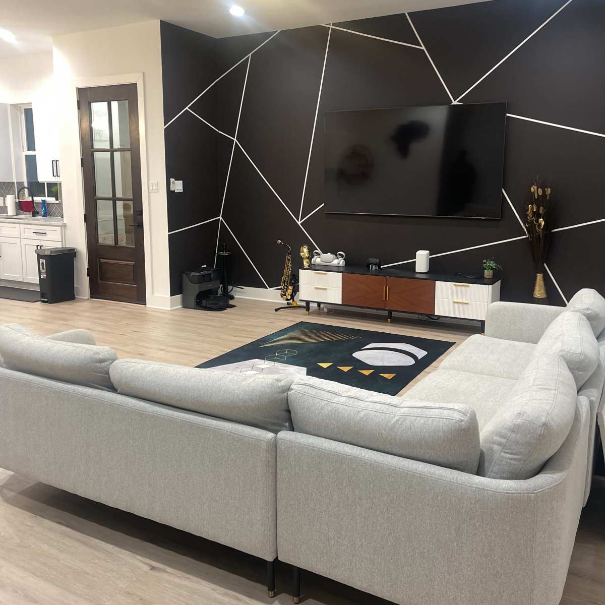 A modern living room features a large sectional sofa facing a wall-mounted television, with a geometric design on the wall and a decorative area rug on the floor.