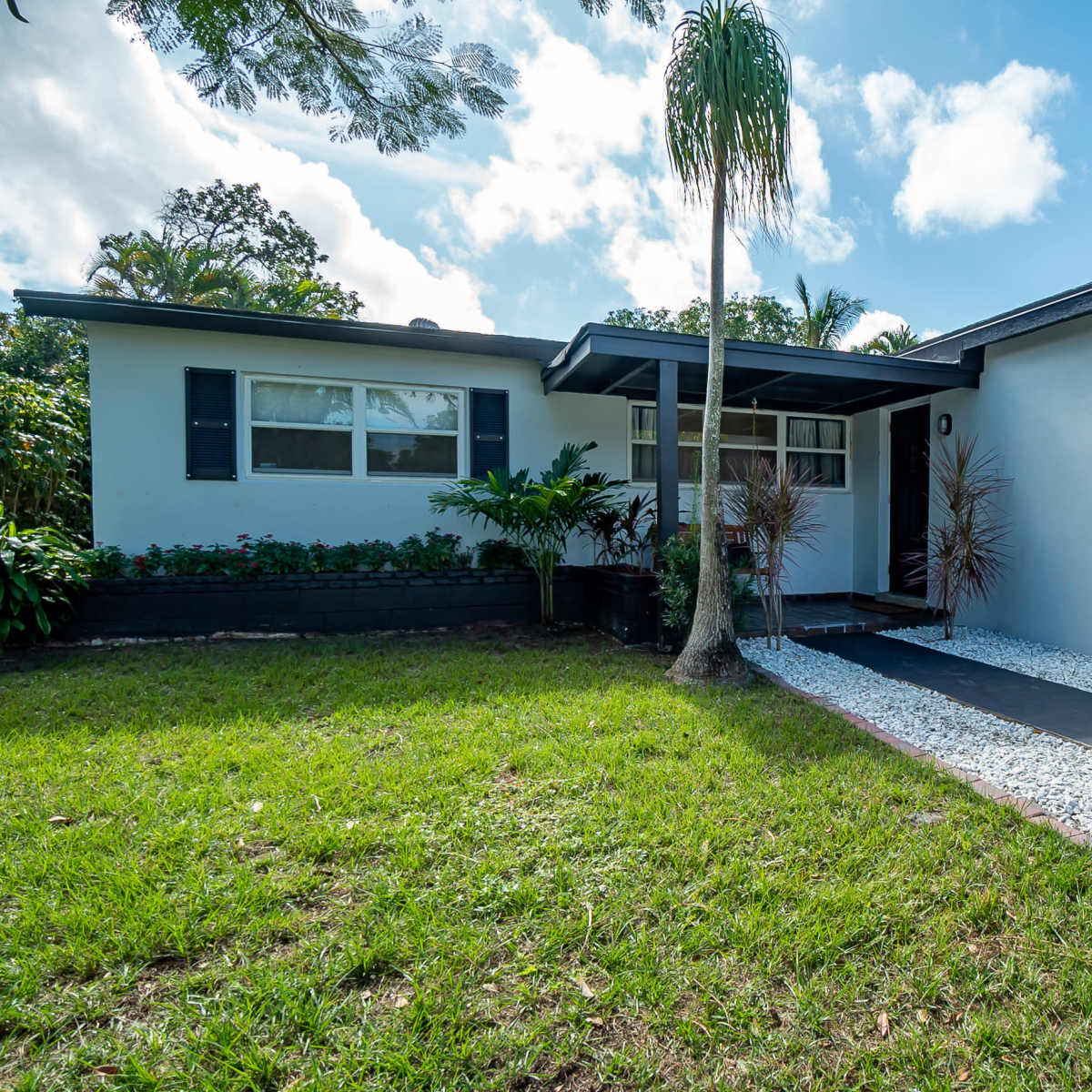 A white house with black shutters and a landscaped front yard featuring a stone pathway and tropical plants.
