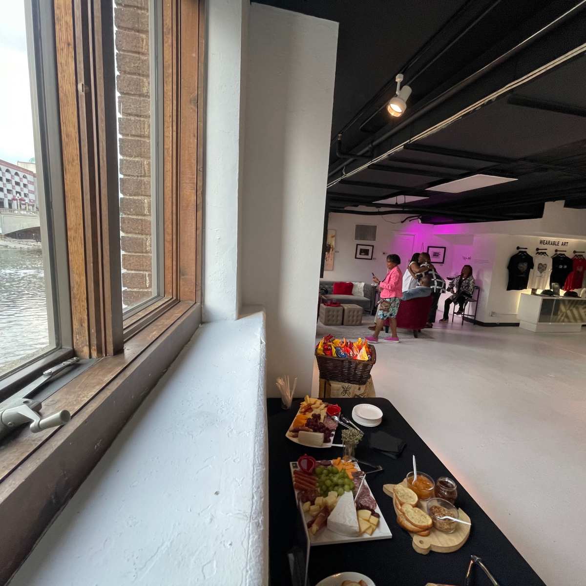 A modern interior space with a table of food in the foreground and people mingling in the background by a large window overlooking a river.