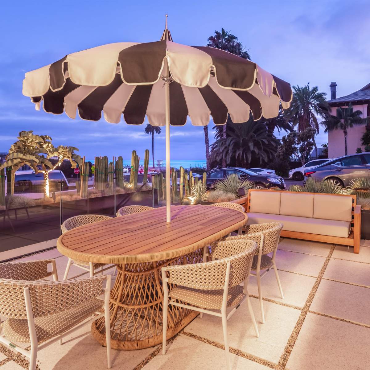 A round wooden table with woven chairs is set under a striped umbrella on a patio surrounded by palm trees and a view of cars parked nearby.