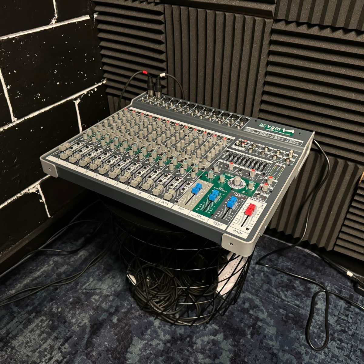 A mixer console is placed on top of a wire basket in a soundproofed room with textured black walls.