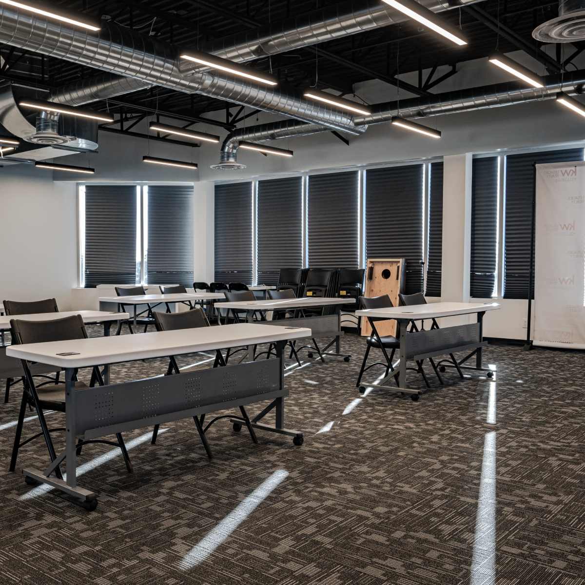 A modern classroom features several folding tables and chairs arranged under bright overhead lights, with large windows partially covered by shades.