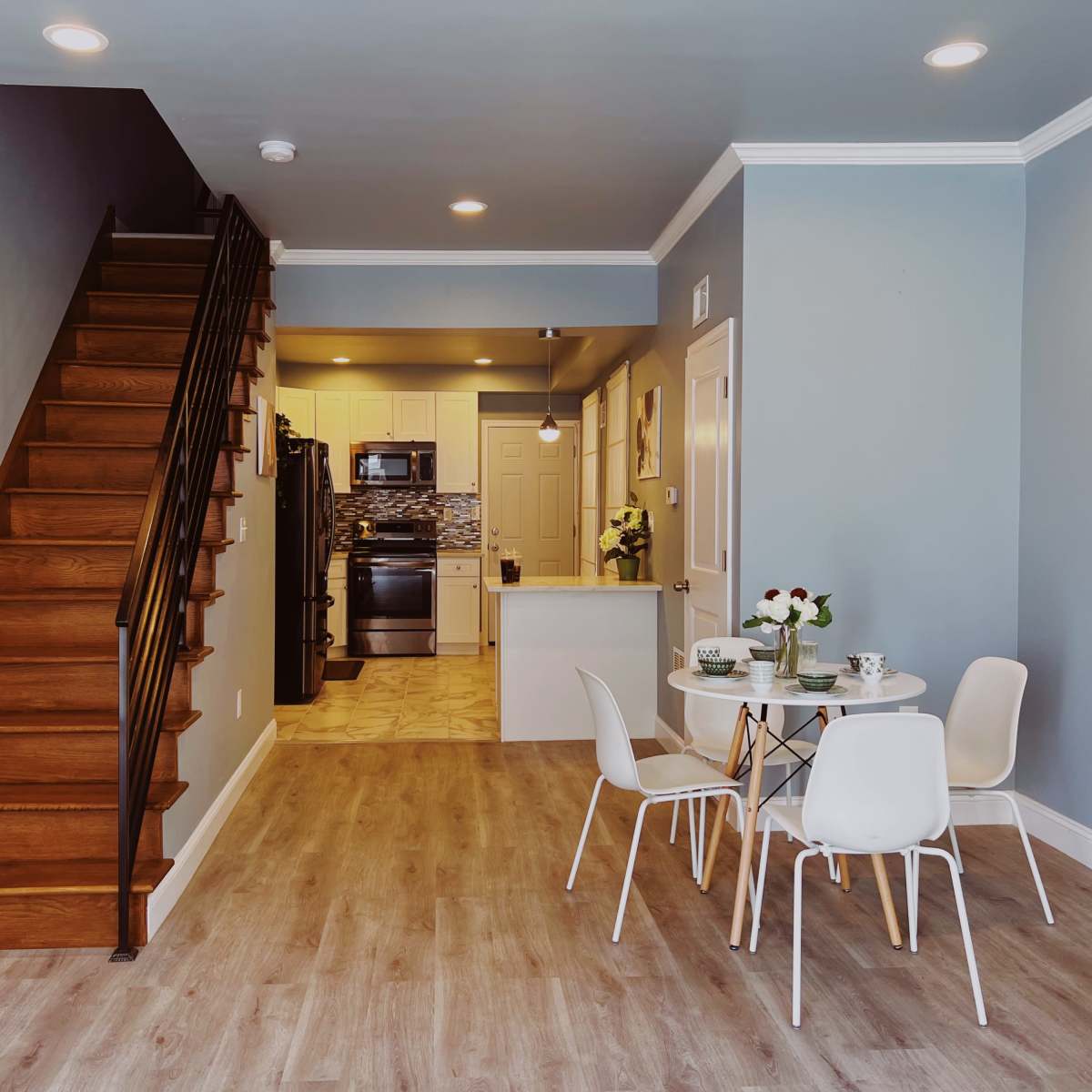 A modern living space features a staircase leading to the upper level, a kitchen visible in the background, and a small dining area with a round table and chairs.