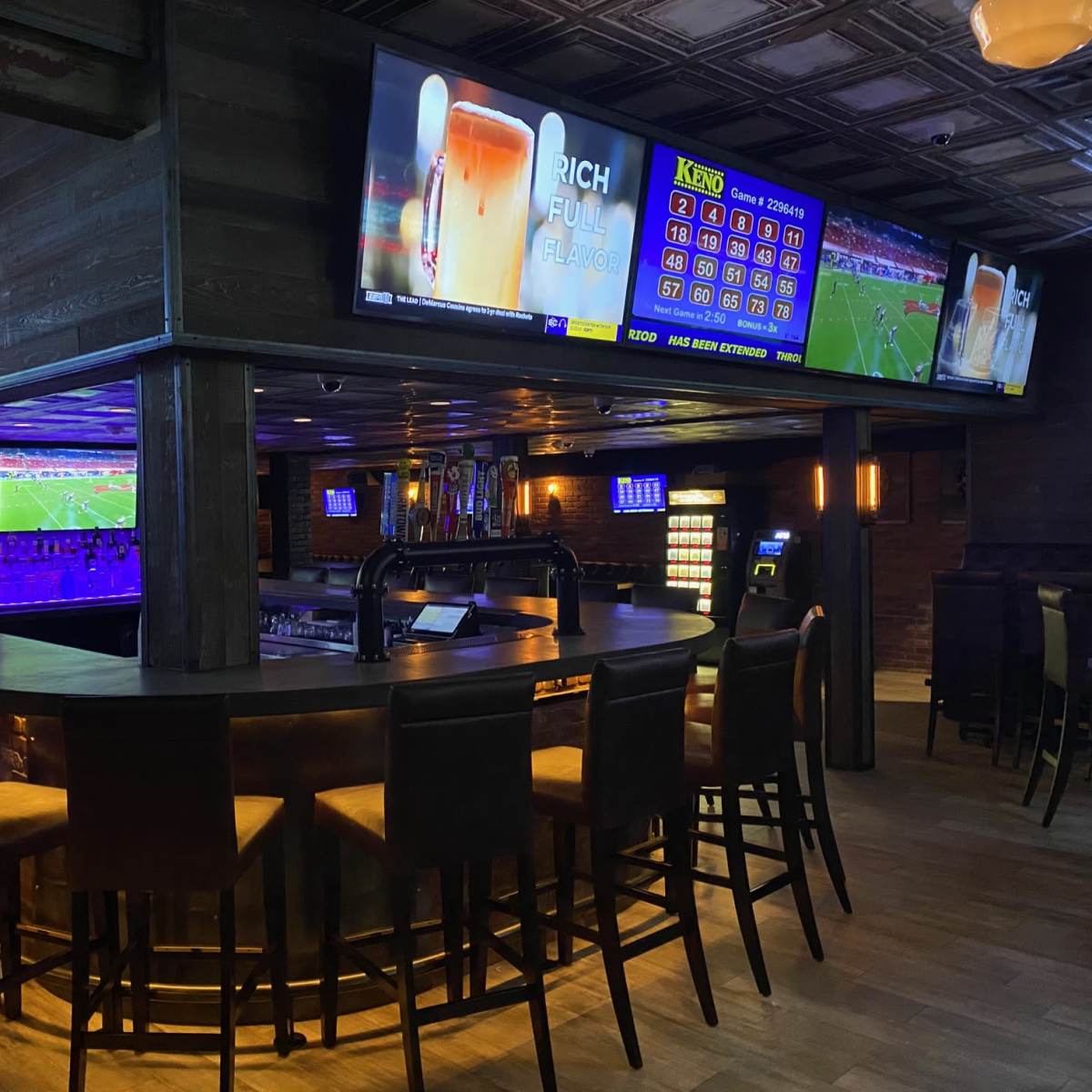 A dimly lit bar with high stools, multiple large screens displaying sports events, and a curved counter lined with bottles.