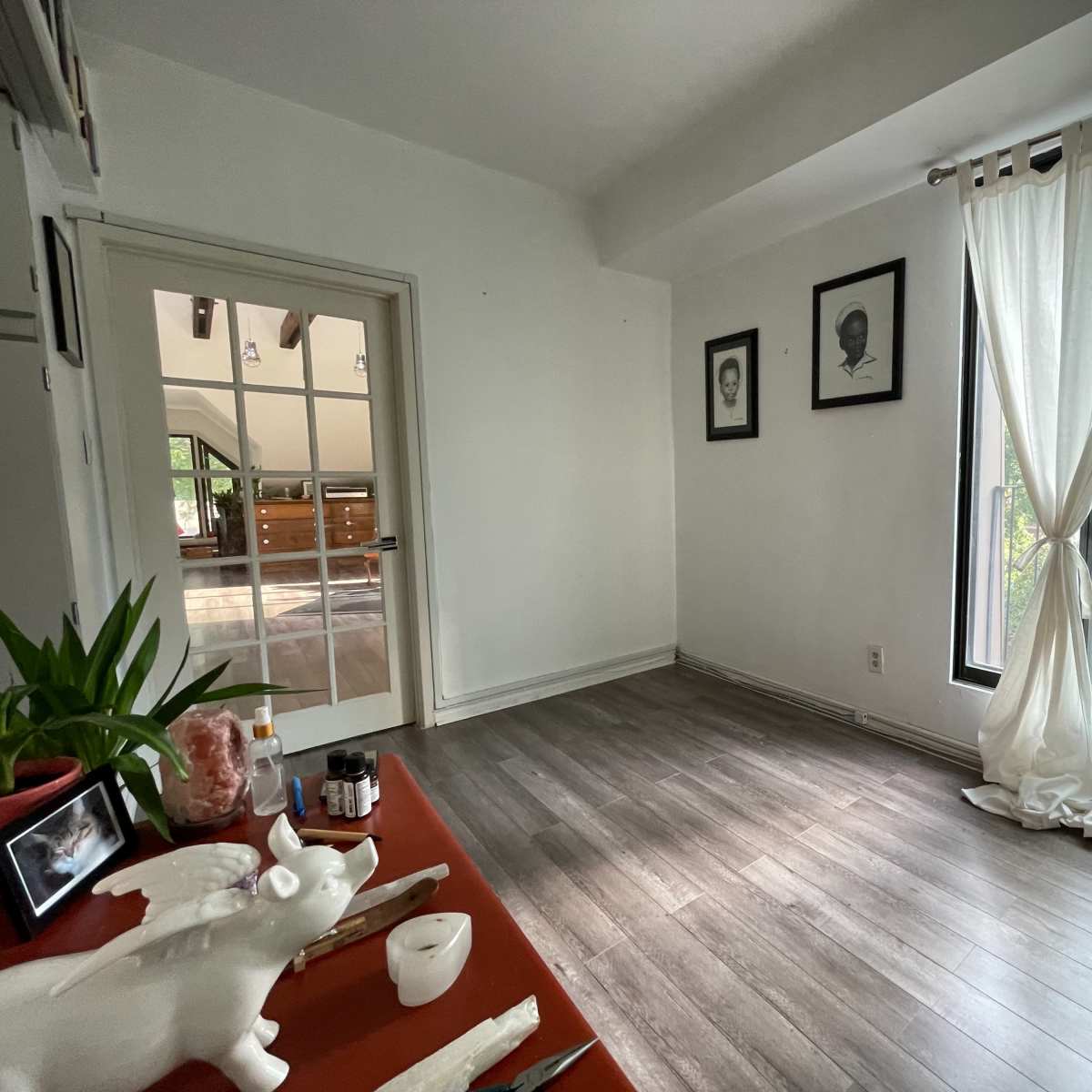 A bright room features large windows with sheer white curtains, hardwood flooring, and a small table with decorative items.