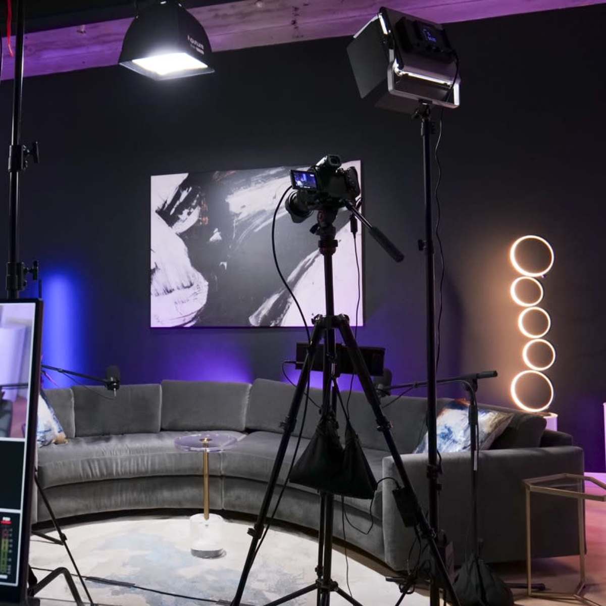 A studio setup for video production featuring a circular gray couch, professional lighting equipment, and a control monitor displaying various camera feeds.