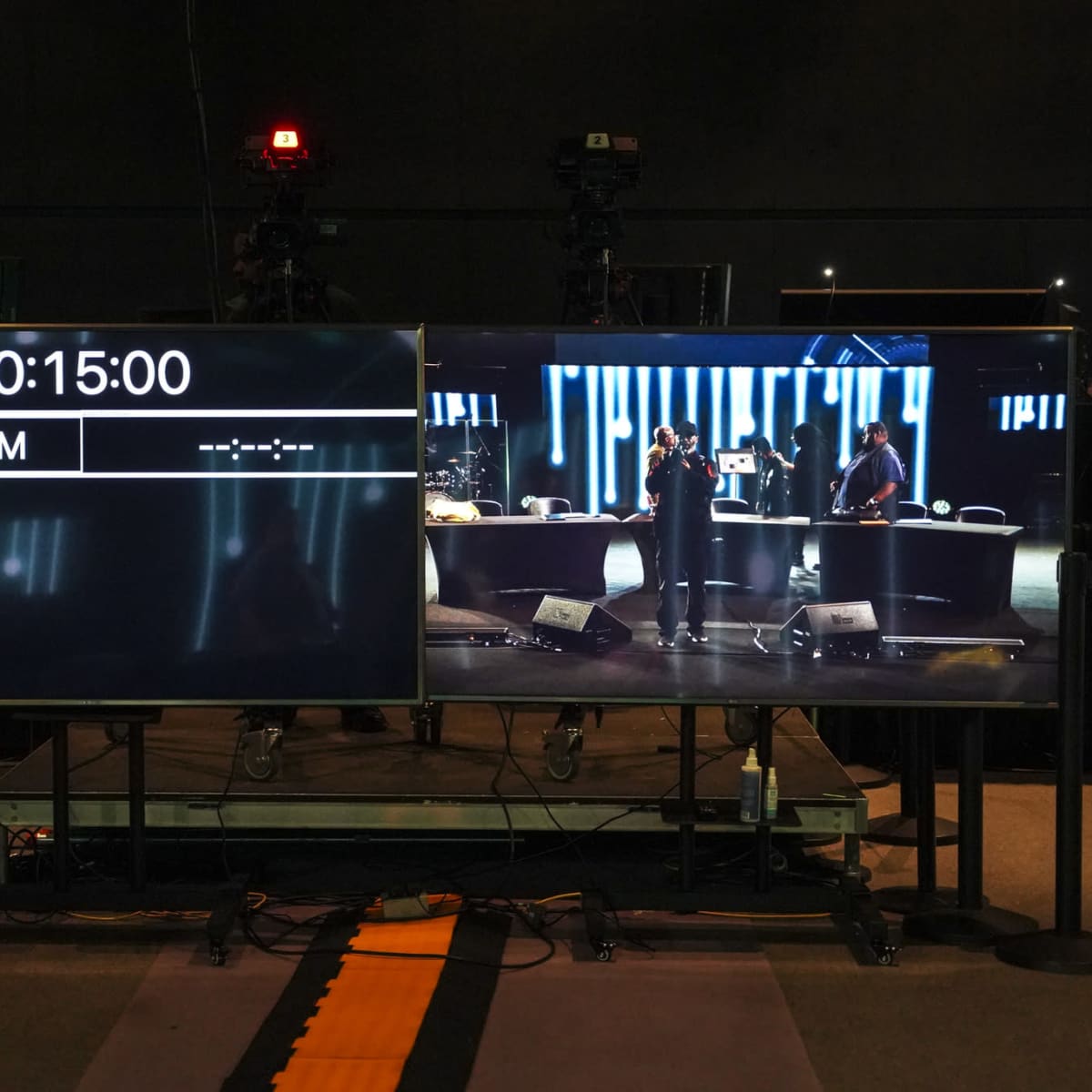 Two large screens in a studio, one displaying a countdown timer and the other showing a live performance on stage.