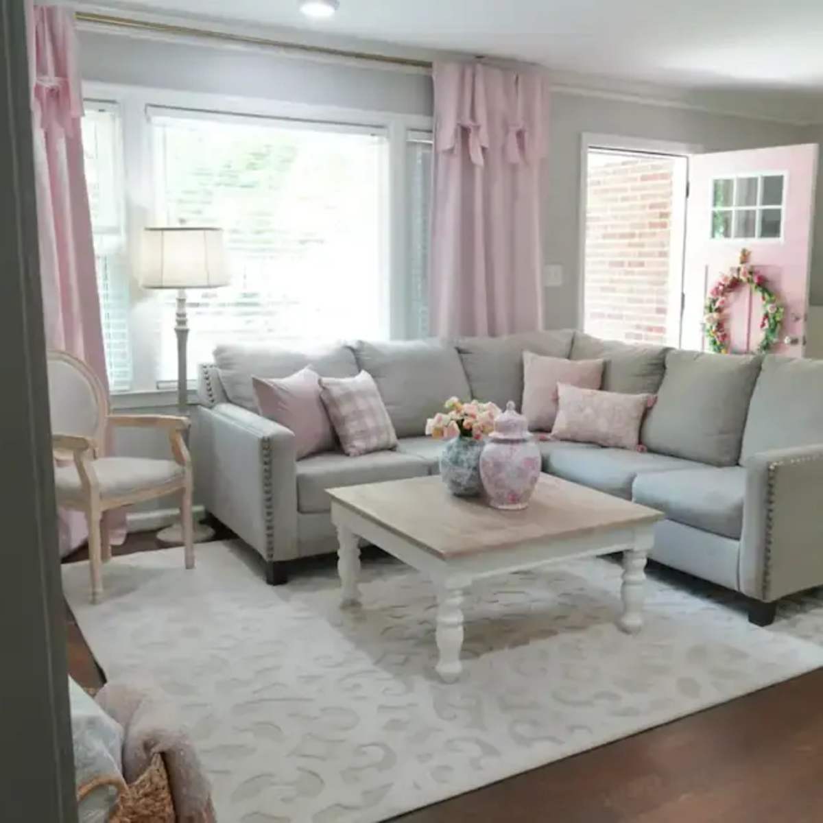A living room features a gray sectional sofa, a white coffee table, and pastel-colored decor near large windows with pink curtains.