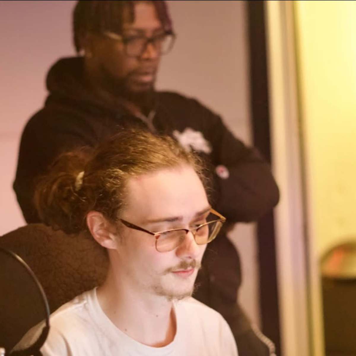 A person with glasses sits at a recording console while another person stands behind them in a studio setting.