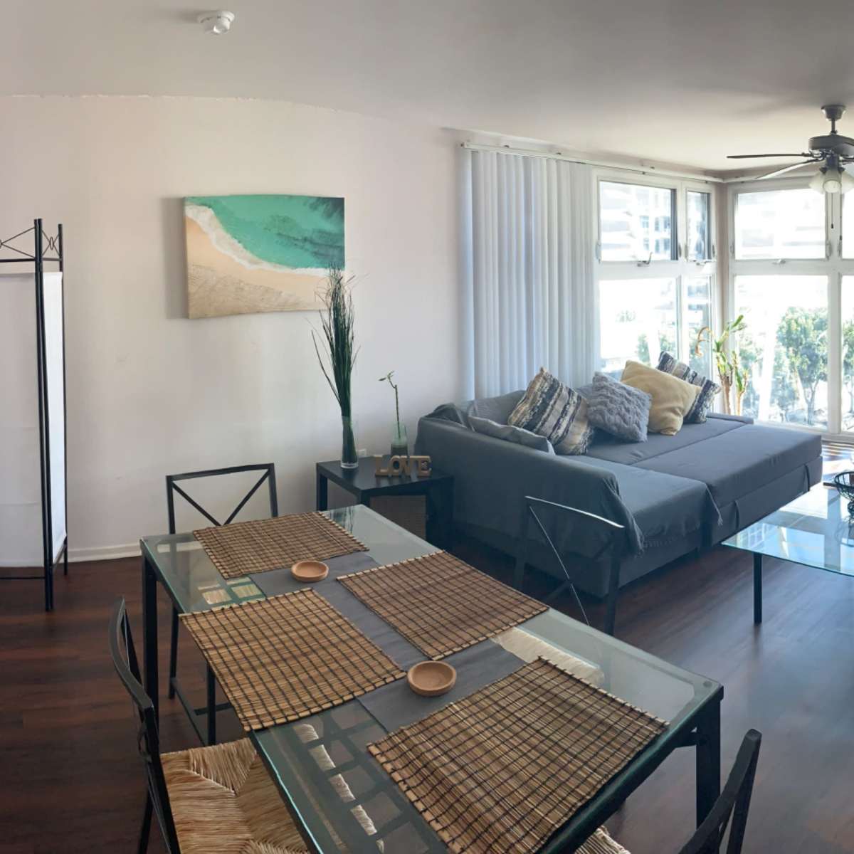 A modern living area with a grey couch, a glass coffee table, a dining table set for four, and large windows offering a view of the outdoors.