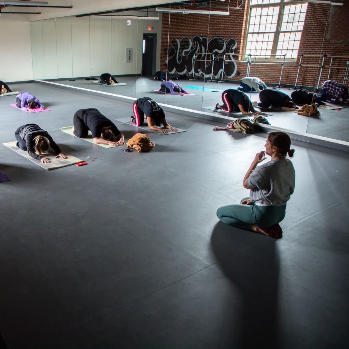 A group of people practice yoga on mats in a spacious studio with large mirrors and exposed brick walls.