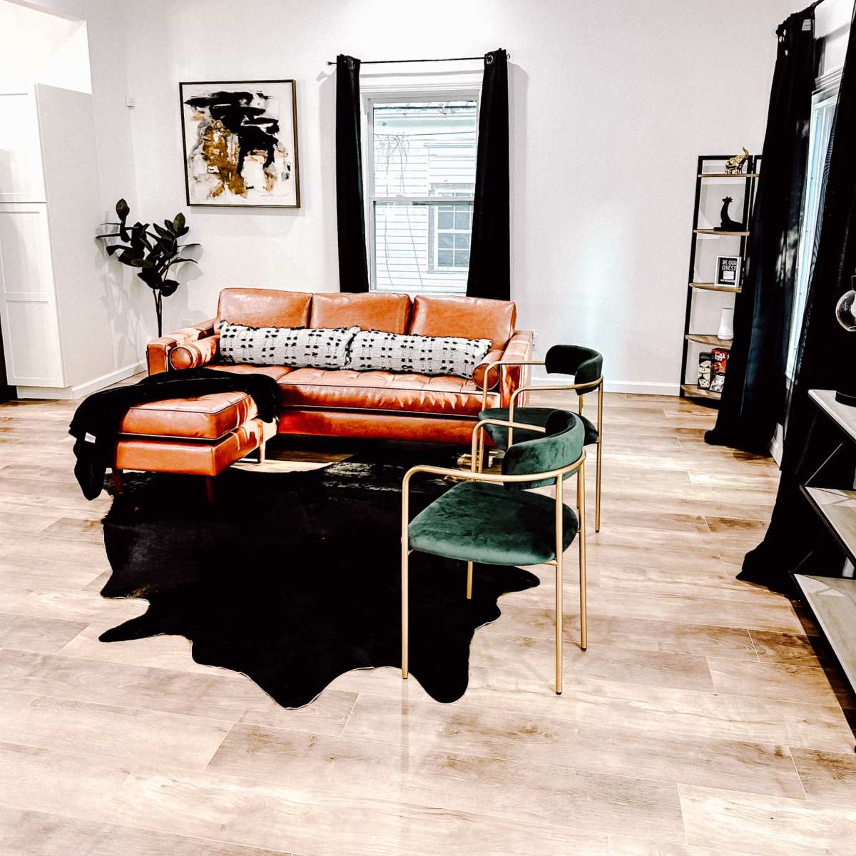 A modern living room with a brown leather sofa, two green chairs, a black rug, and large windows draped with black curtains.