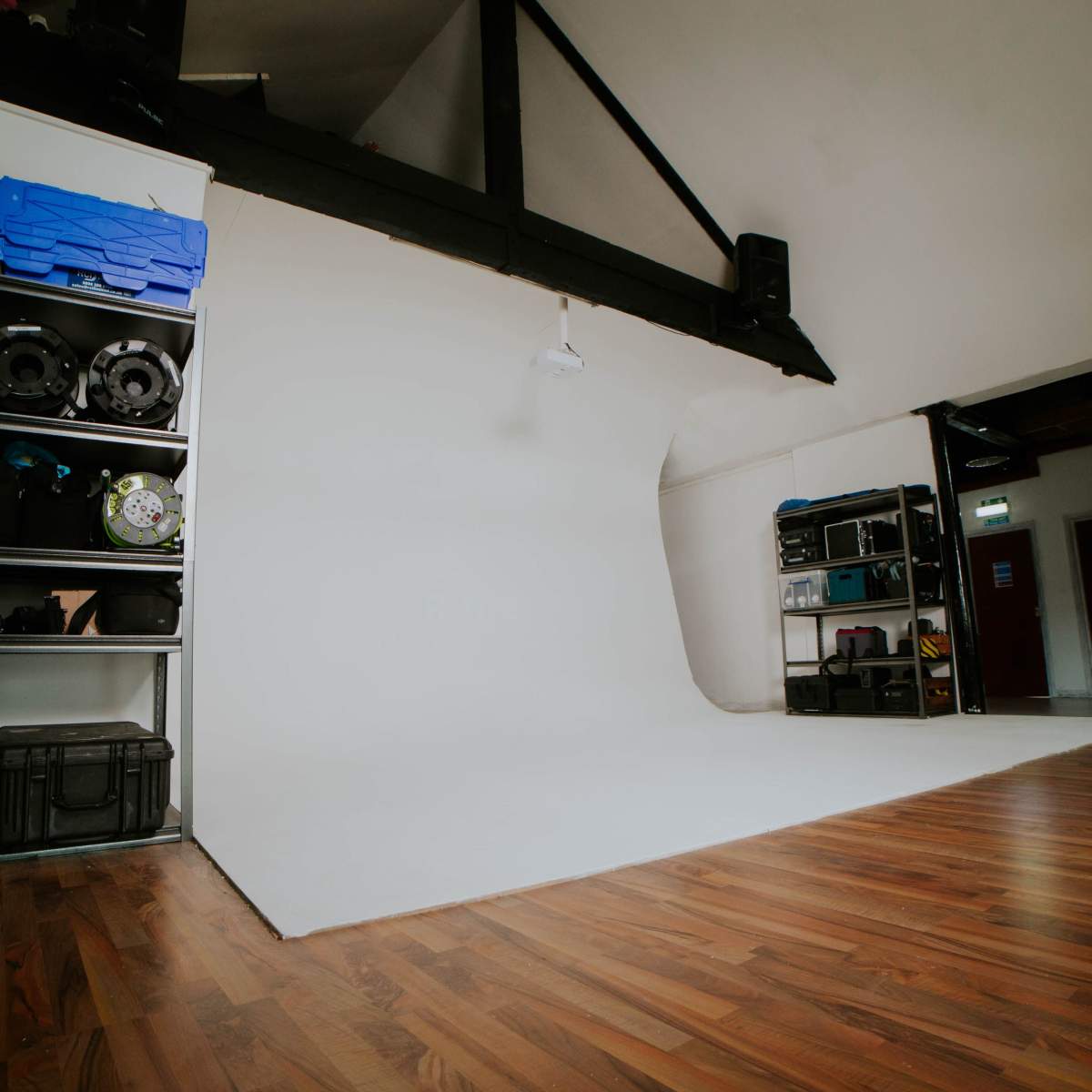 A photography studio with a white backdrop, wooden flooring, and shelves containing various equipment on one side.