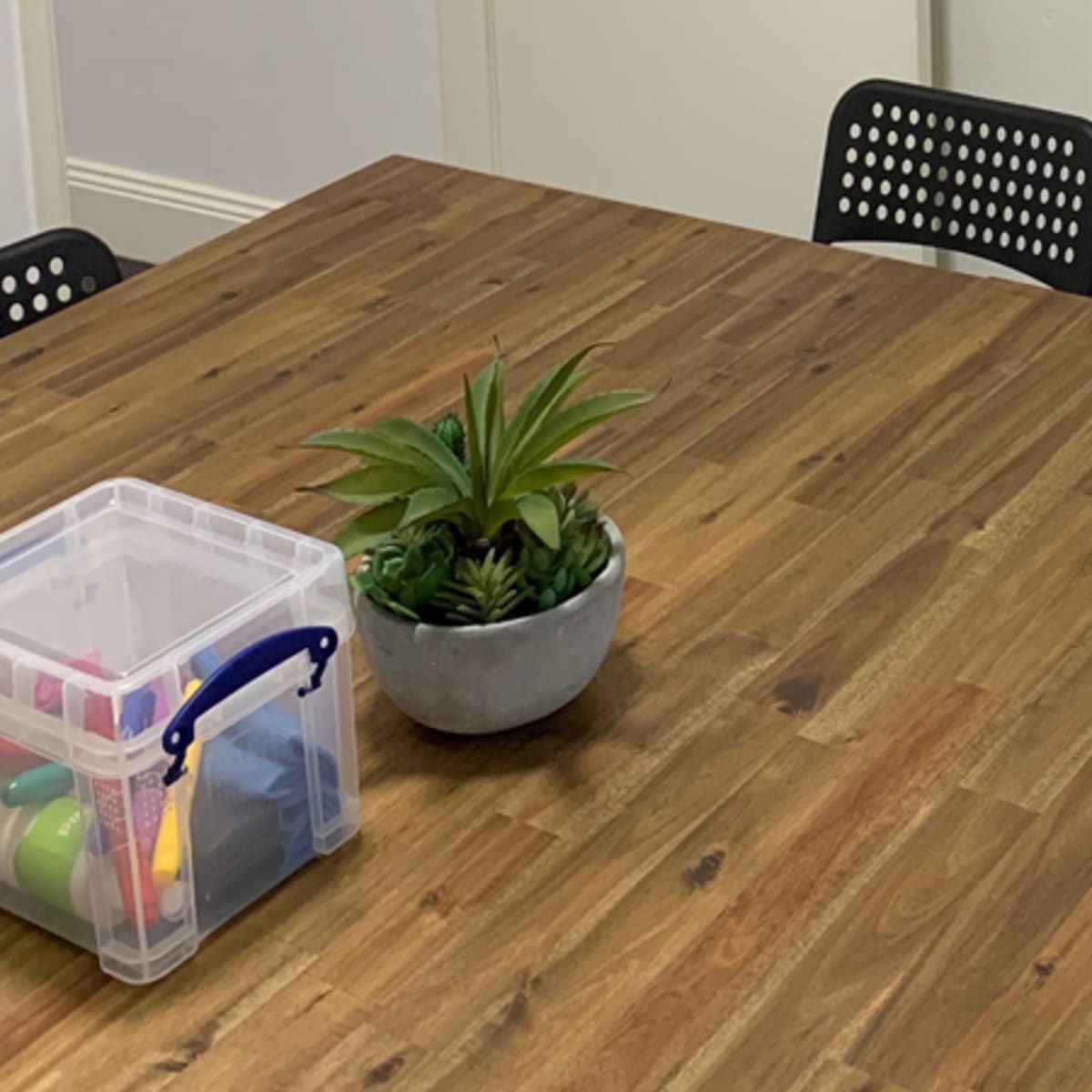 A wooden table with a decorative plant and a clear container filled with colorful items is surrounded by four black chairs in a plain room.
