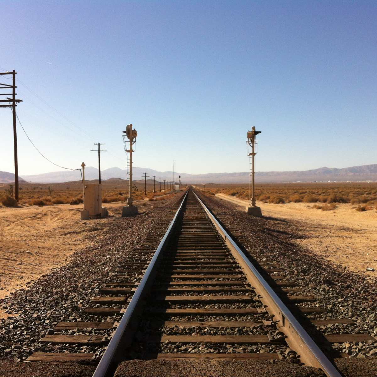 A straight railway track stretching through a desert landscape with telegraph poles and signal lights on either side.