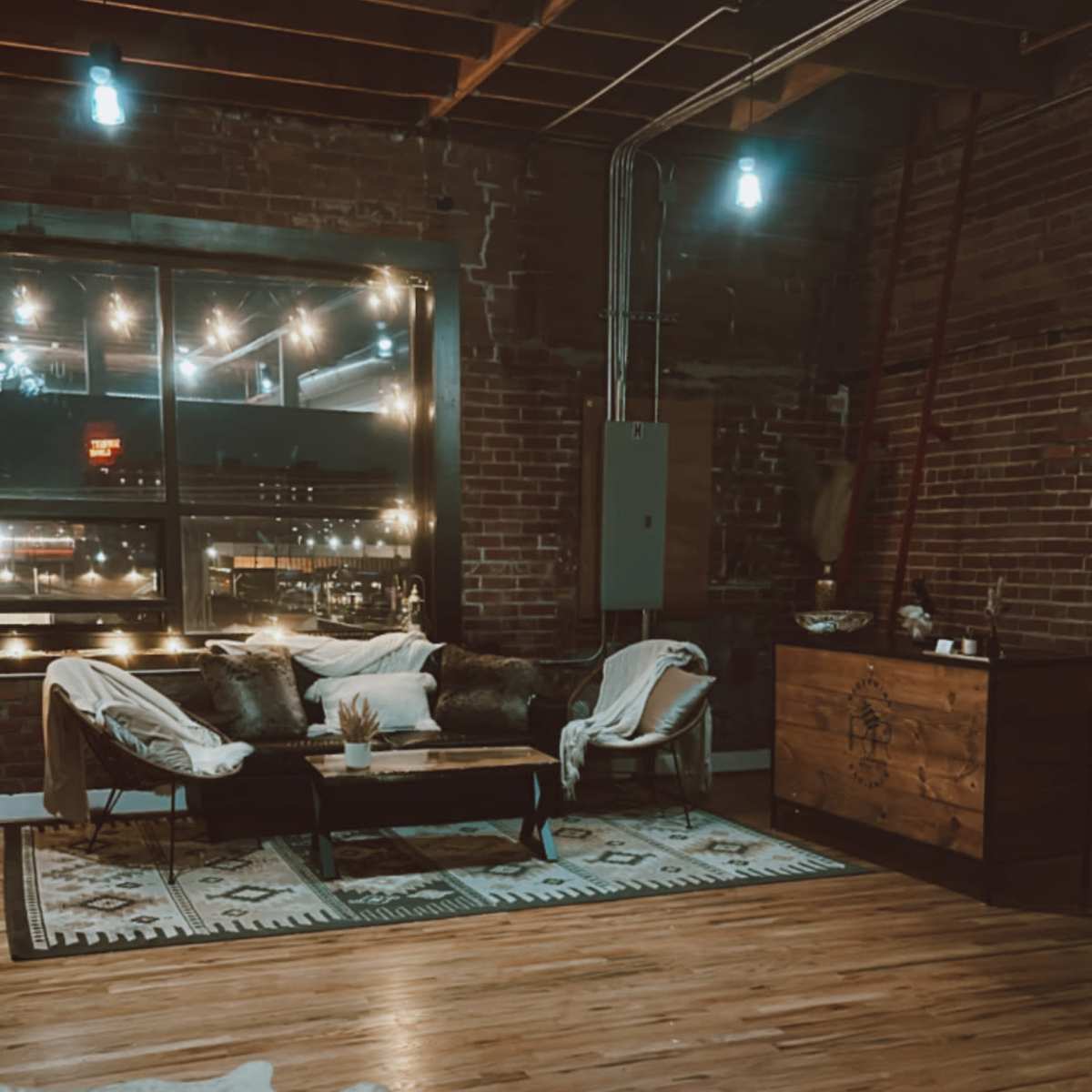 A spacious living area features a wooden floor, a couch and chairs with throw pillows, a patterned rug, a wooden cabinet, and large windows illuminated by string lights.