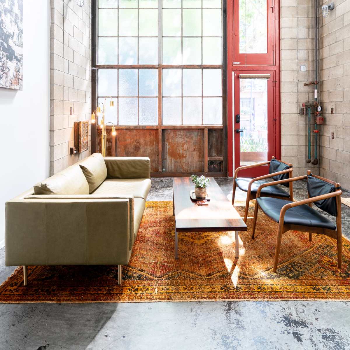 A modern living room with a large window, a green sofa, two black chairs, a coffee table, and a colorful rug on a concrete floor.