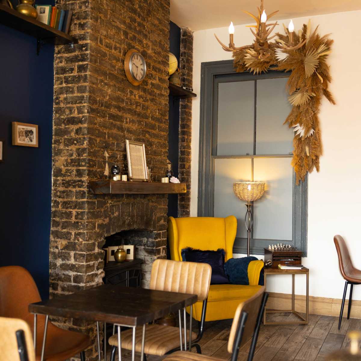 A cozy café interior with a yellow armchair, wooden tables, and a decorative wall featuring a large arrangement of dried plants above a fireplace.