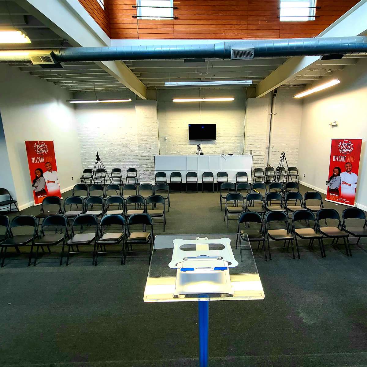 A conference room set up with rows of chairs facing a stage, flanked by two promotional banners and equipped with cameras and a monitor.