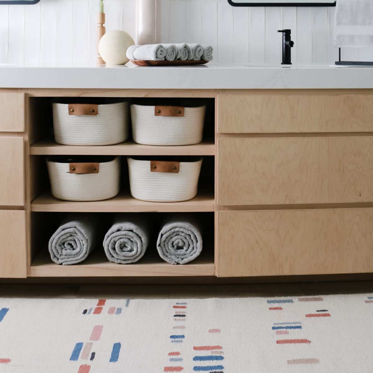 A modern bathroom features a wooden vanity with wicker baskets and neatly rolled towels beneath mirrors, alongside decorative pampas grass in a vase.
