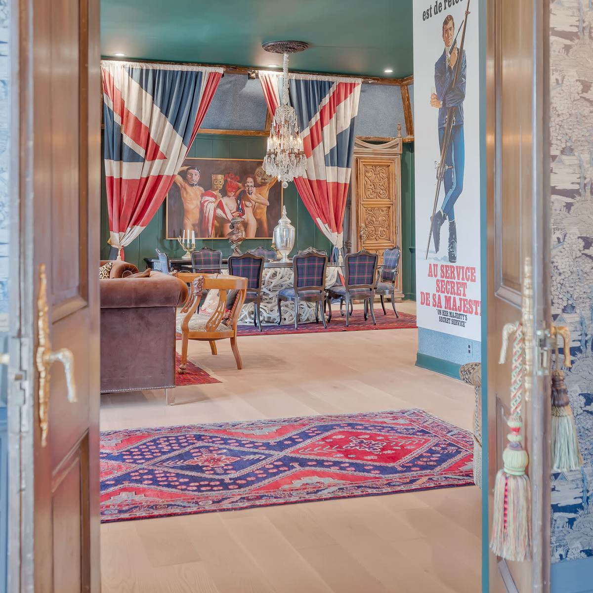 A doorway opens into a room featuring Union Jack curtains, a chandelier, and a mural of a character in suit against patterned wallpaper.