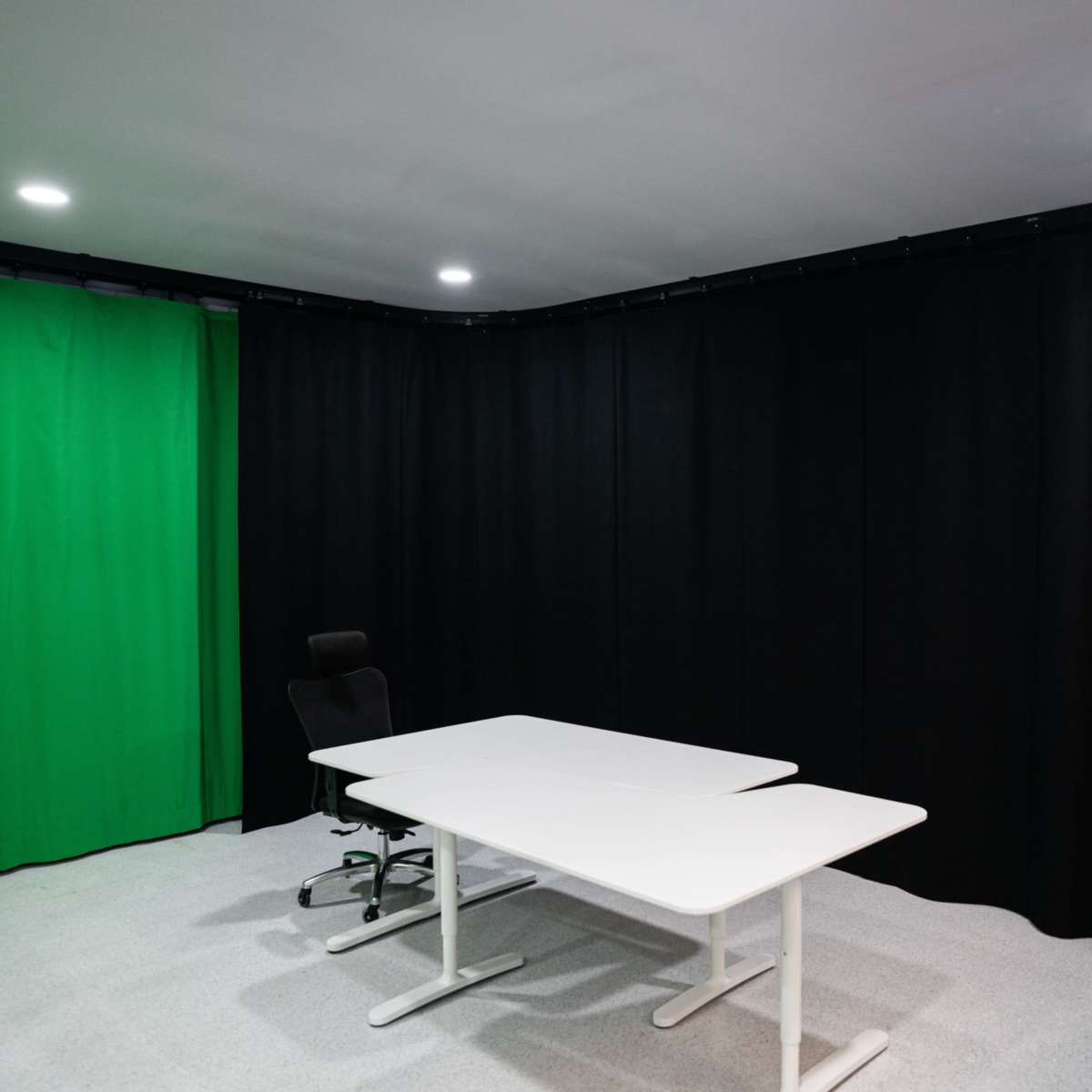 A small studio room features a green screen backdrop, black curtains, a white table, and an office chair.