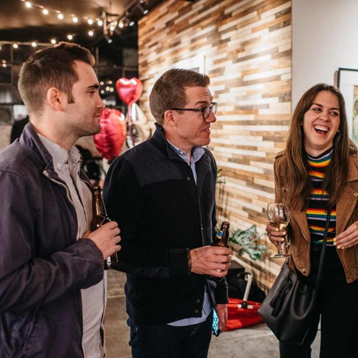 Four people are engaged in conversation at a social gathering, with two holding drinks and wearing casual attire in a warmly lit setting.