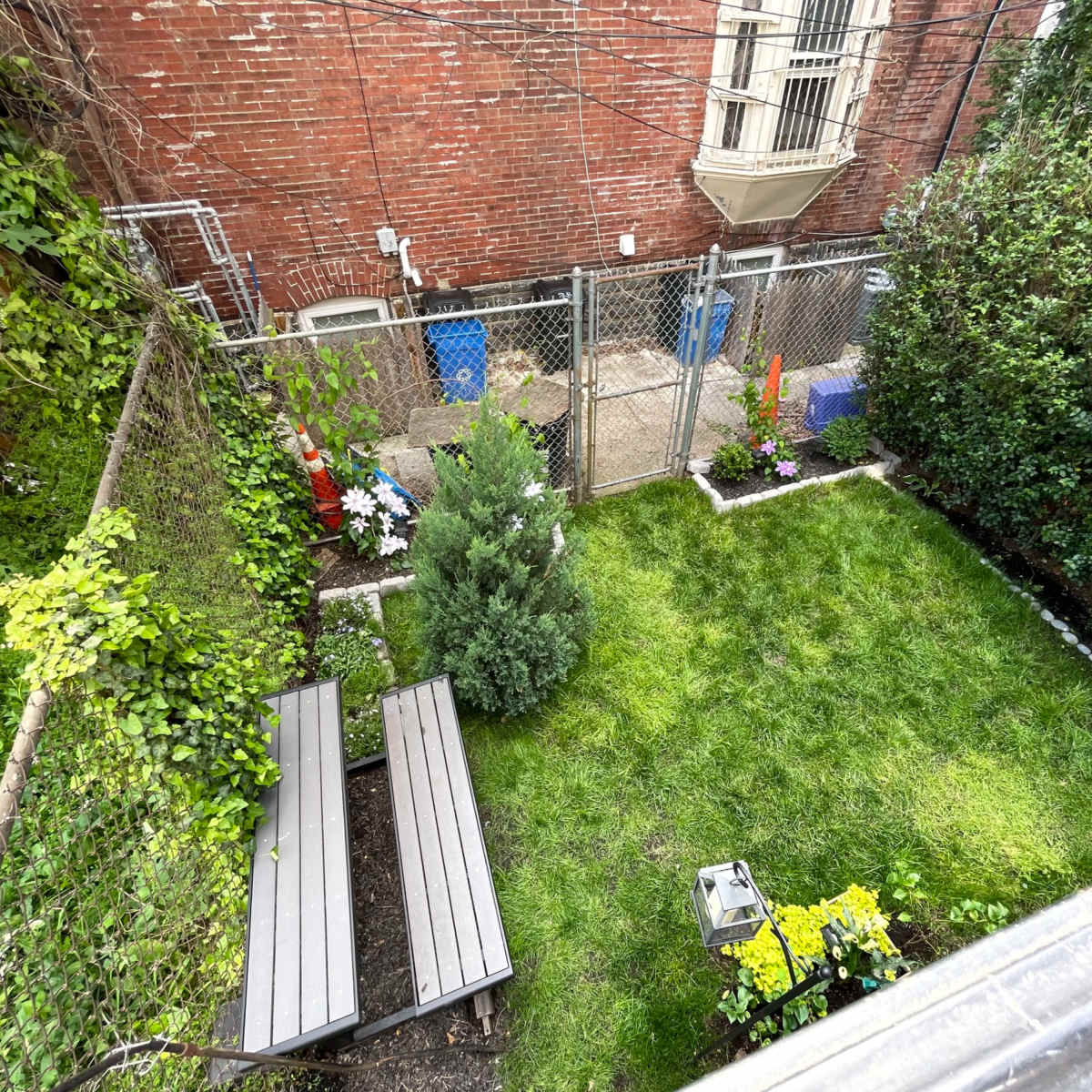 A fenced backyard with a manicured lawn, contains two wooden benches, and is bordered by shrubs and flowers.