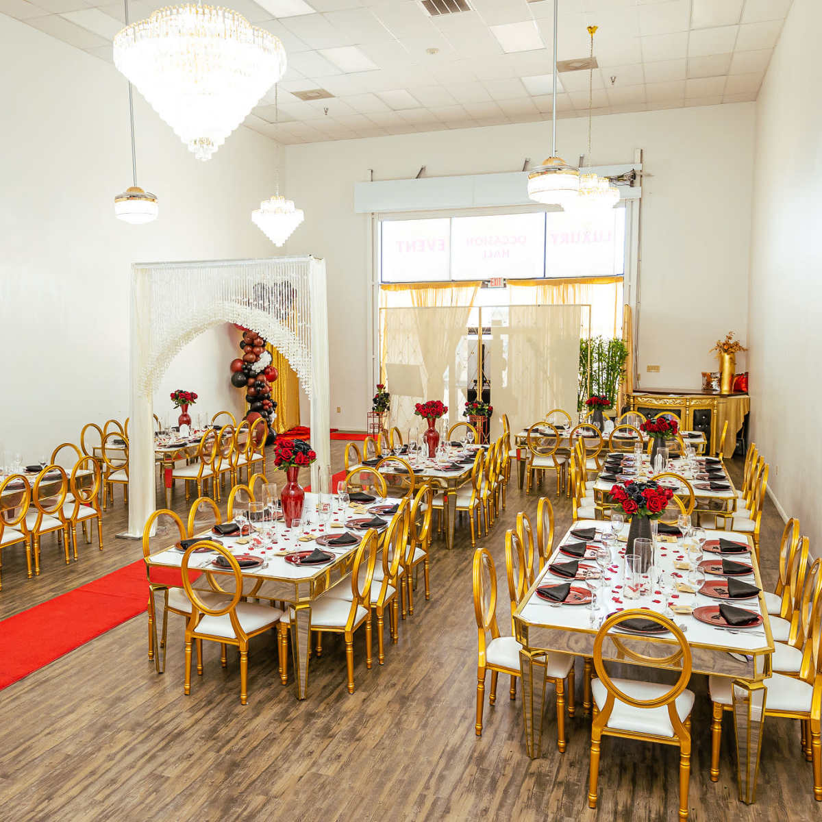 An elegantly arranged dining hall with tables set for an event, featuring gold chairs and red floral centerpieces.