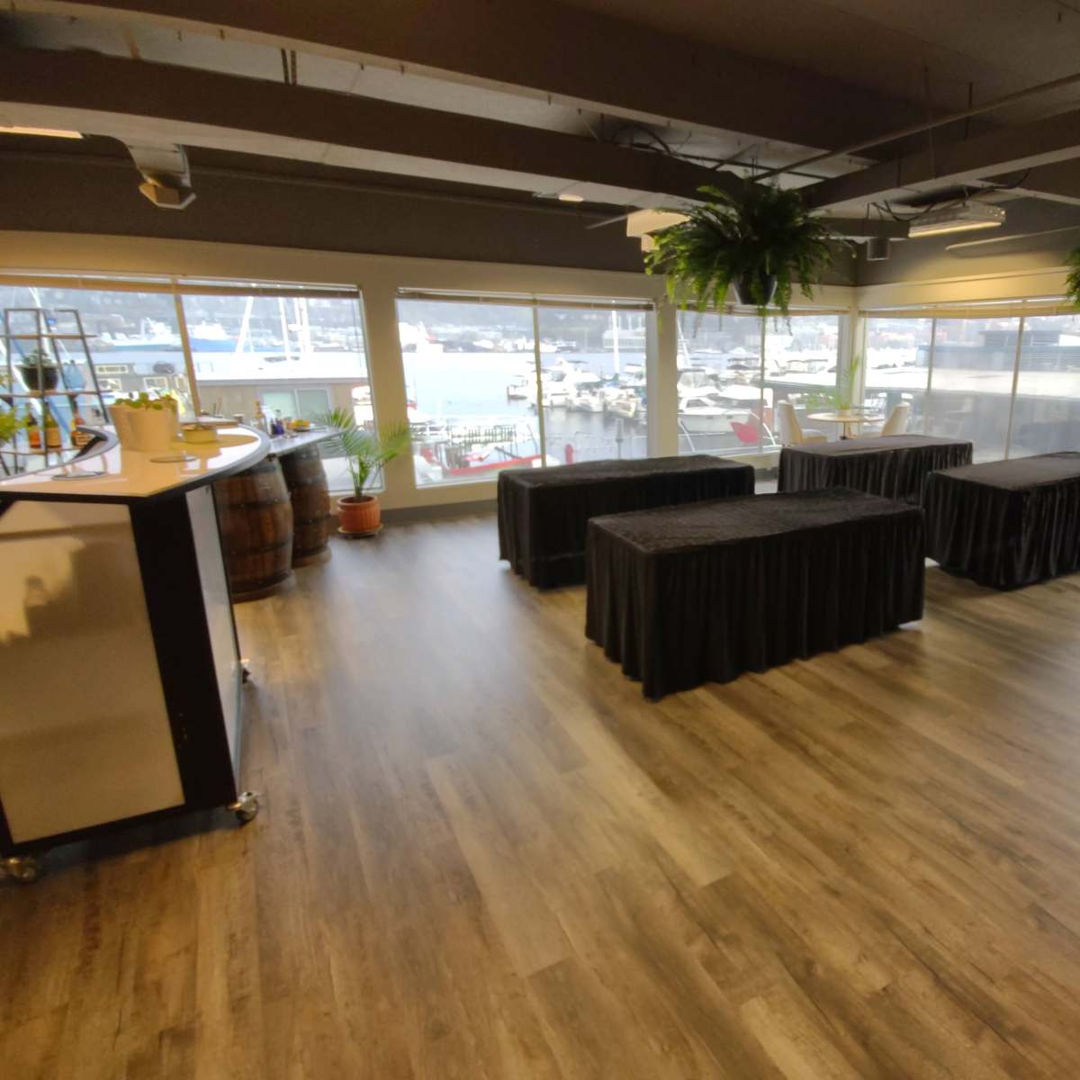 The interior space features several tables covered with black tablecloths, large windows showcasing a view of boats outside, and a bar area with decorative plants.