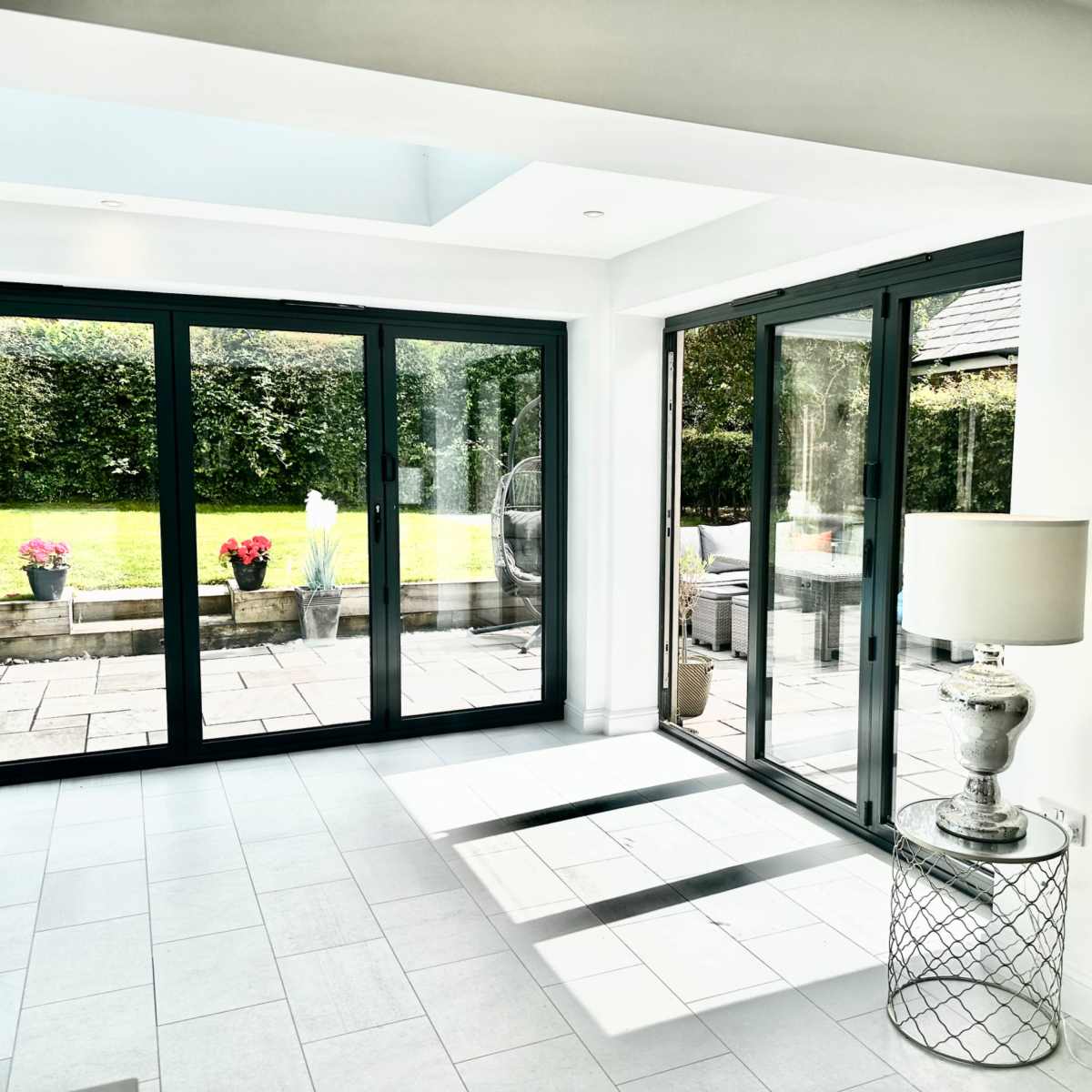 A bright room with large glass doors leading to a garden, featuring a lamp and a decorative table in the foreground.