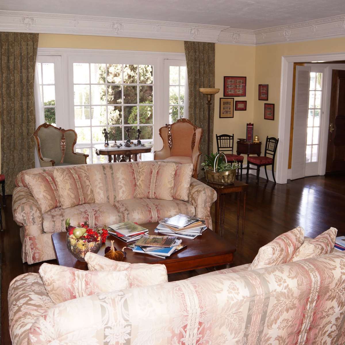 A spacious living room with two patterned couches, a wooden coffee table cluttered with magazines, and large windows letting in natural light.
