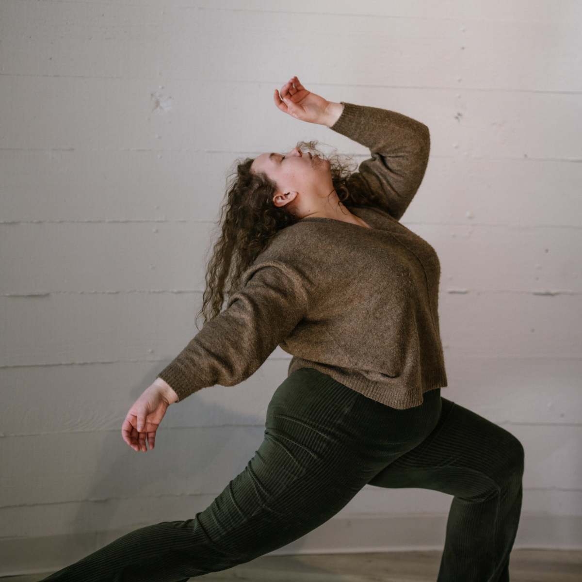 A person with curly hair performs a dynamic dance pose in a cozy, neutral-toned outfit against a textured wall.