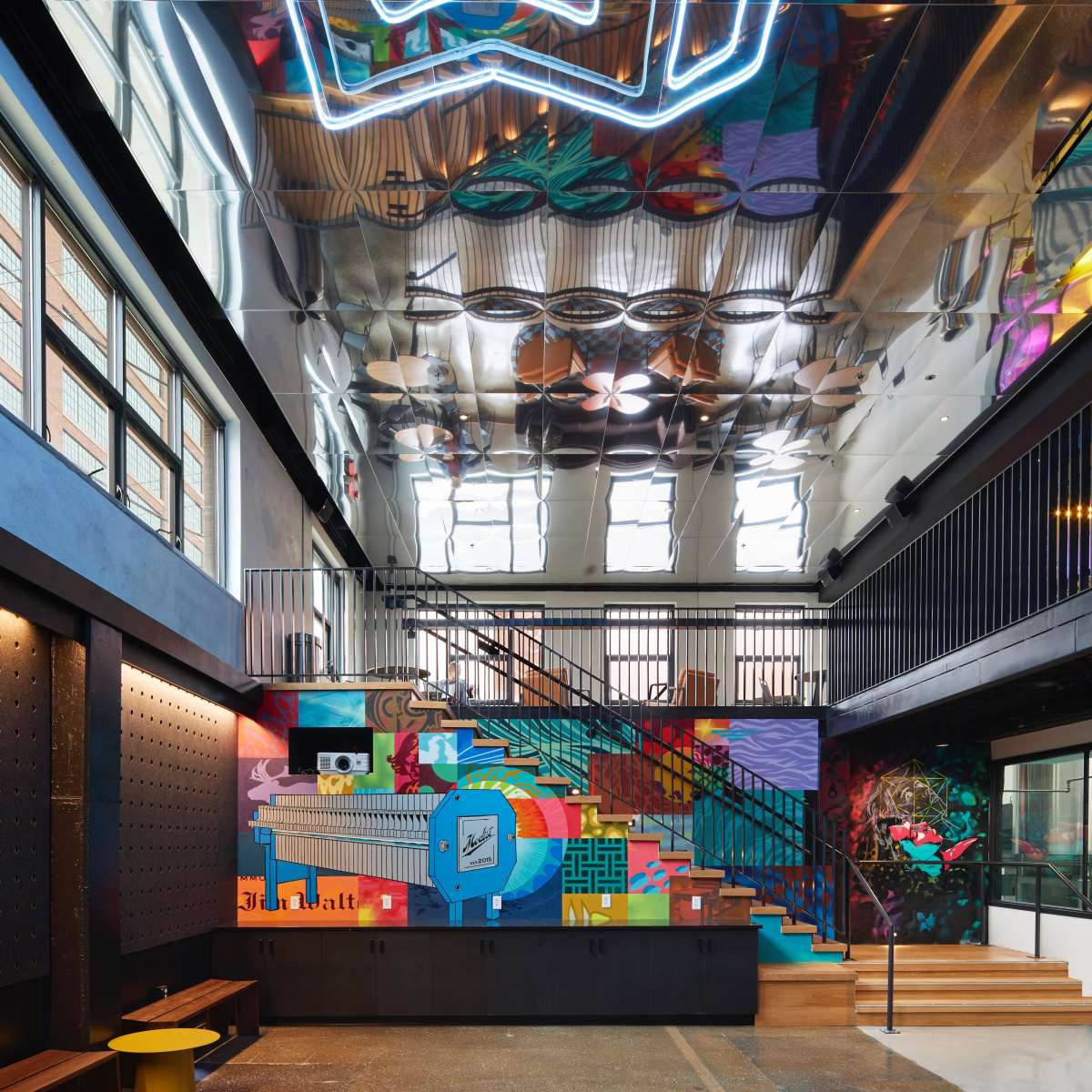 A vibrant interior space featuring a colorful mural, reflective ceiling, and a staircase leading to an upper level.
