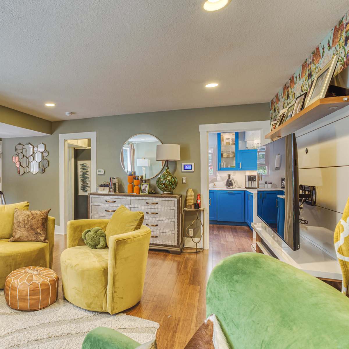 A brightly colored living room features green furniture, a circular mirror, and an open view into a blue-themed kitchen.