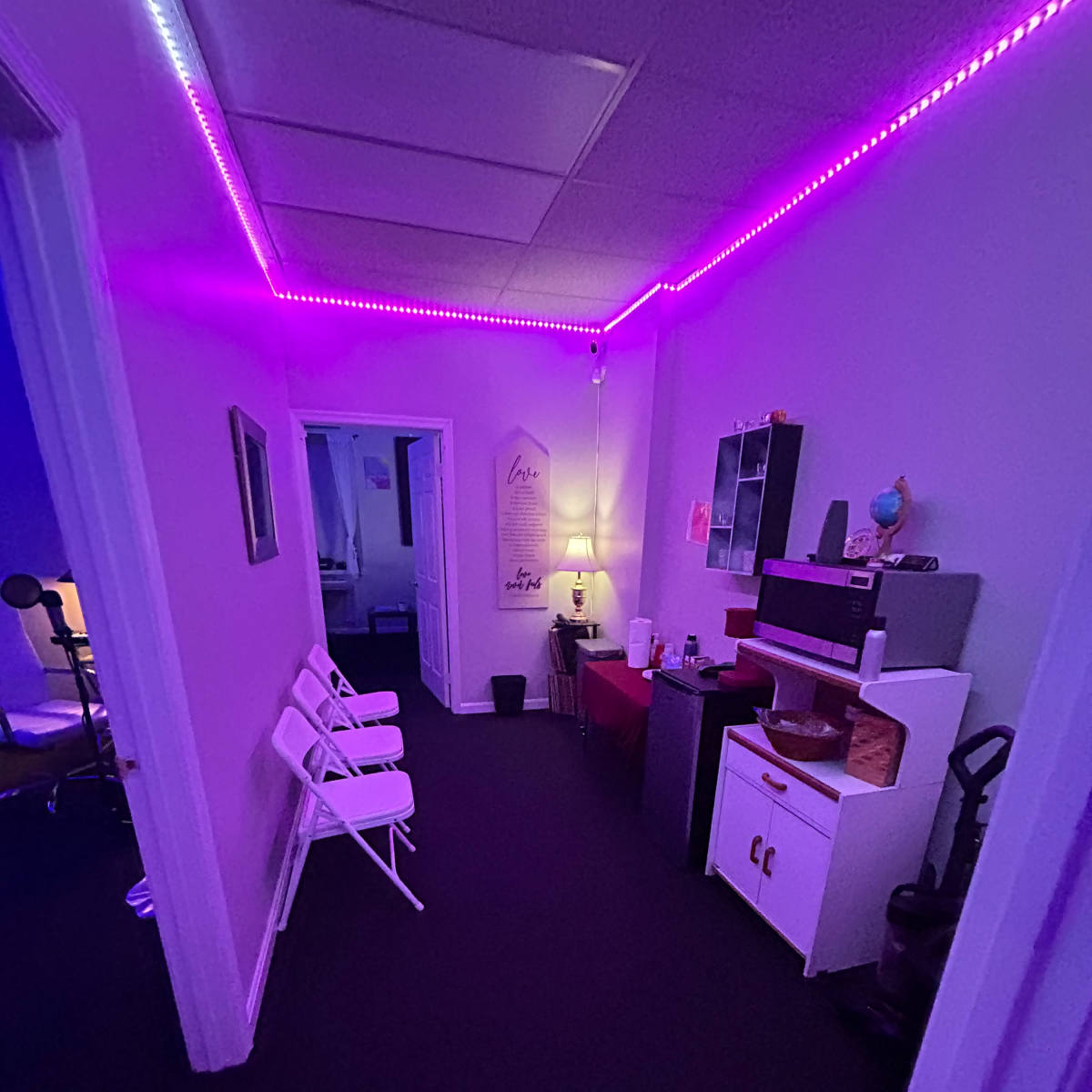 A narrow hallway is lined with purple LED lights, leading to rooms with chairs and various furniture on either side.