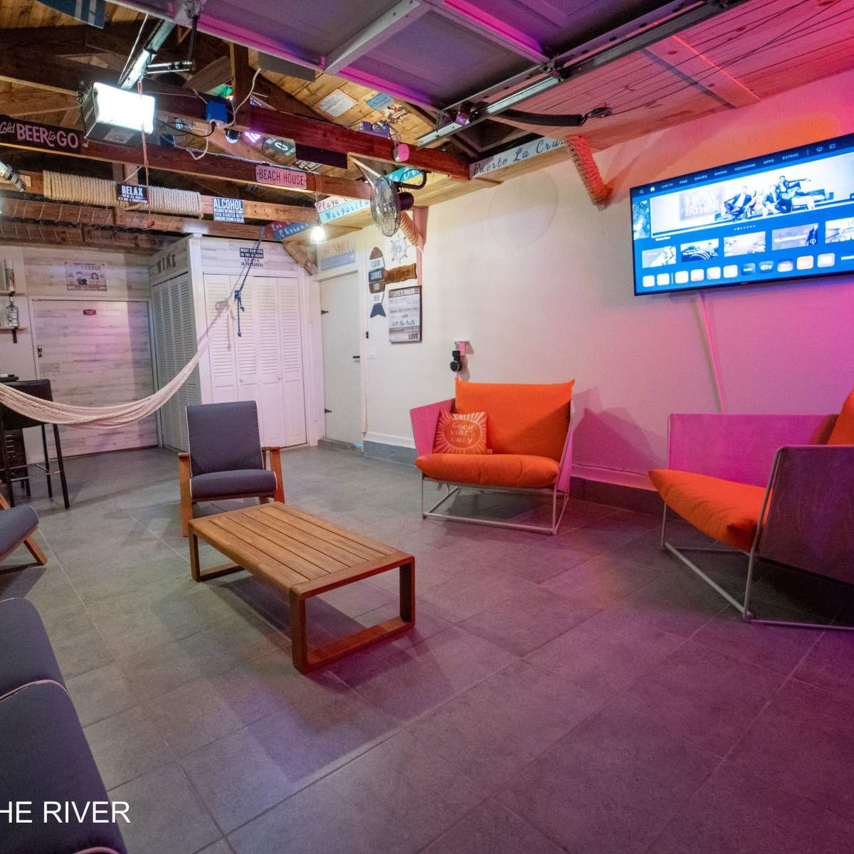A cozy indoor space features lounge seating, a coffee table, a hammock, and a wall-mounted television under a wooden ceiling with exposed beams.