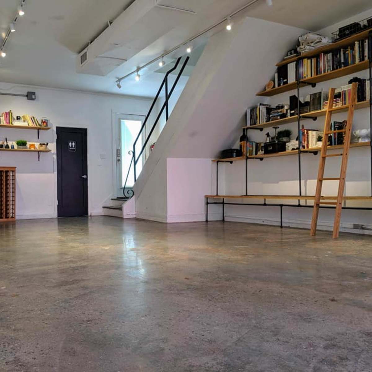 A minimalist interior space featuring polished concrete flooring, a staircase, shelves with books, and a small table near the entrance.