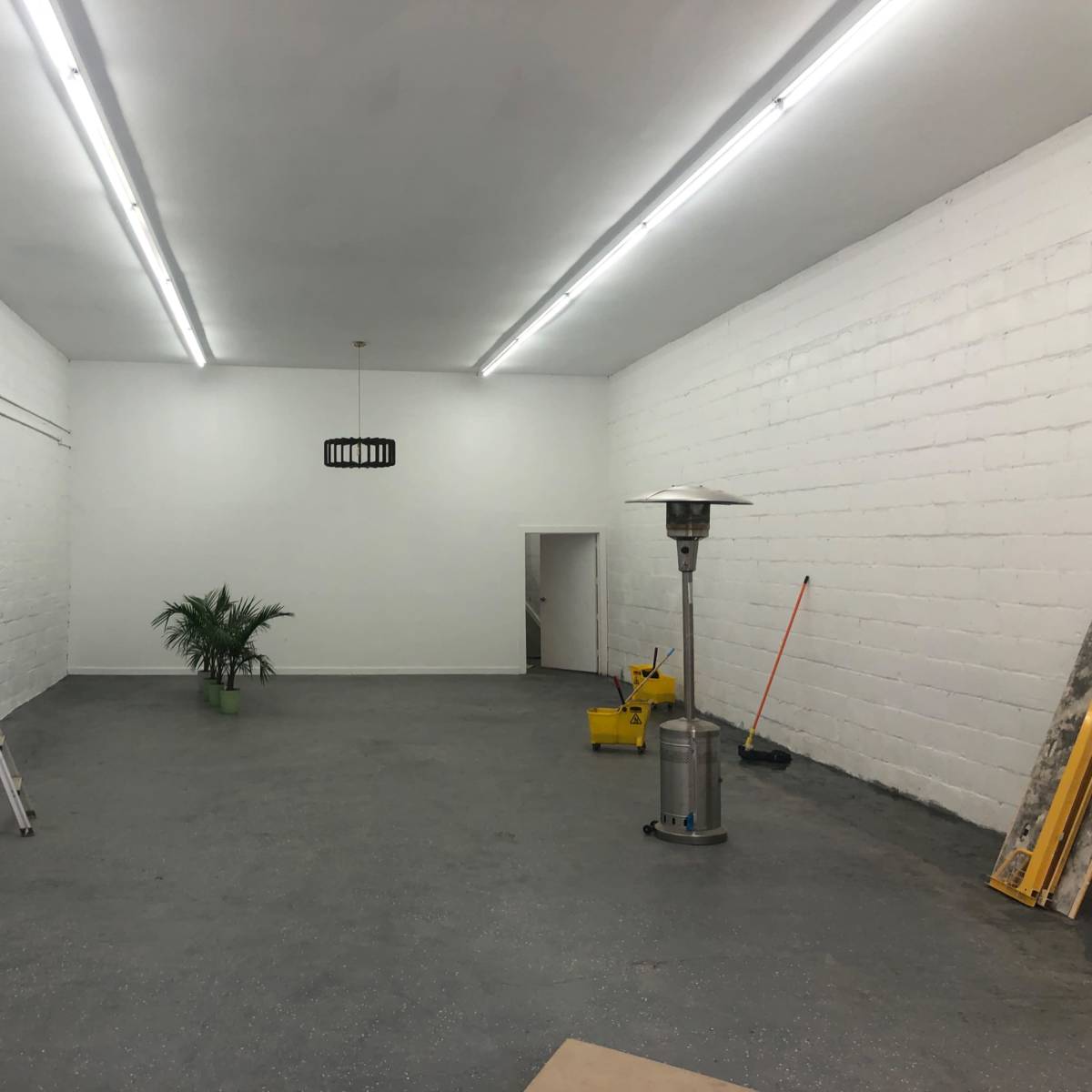 A spacious, empty room with white walls, a plant in a green pot, a ladder, a patio heater, and a door leading to another space.