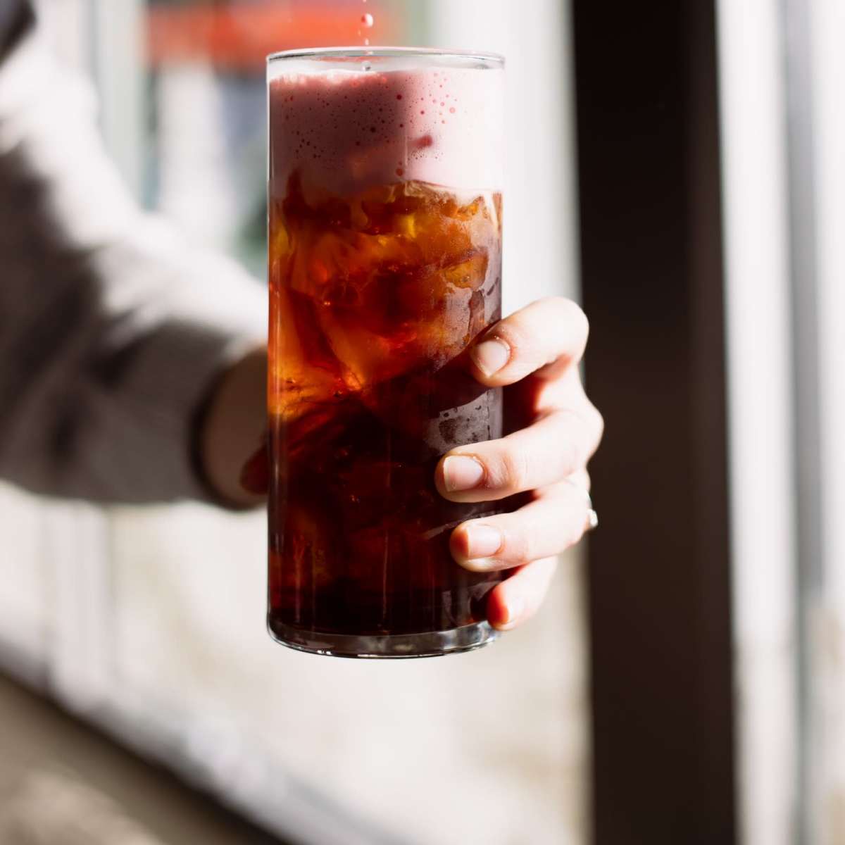 A hand holds a tall glass filled with iced tea or coffee as a drop of syrup drips into it.