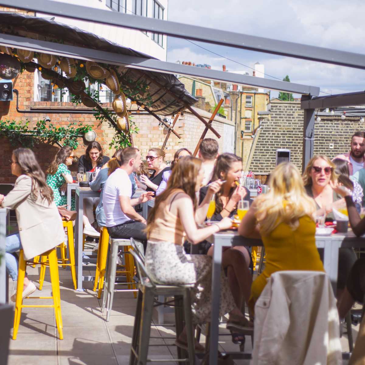 A bustling rooftop bar features groups of people seated at tables, enjoying drinks and conversation under a partially covered area.